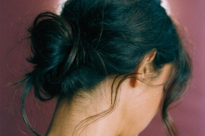 A woman shown from the back and side, wearing a dark messy bun hairstyle with loose strands, set against a dusty mauve background with a soft white halo behind her head.